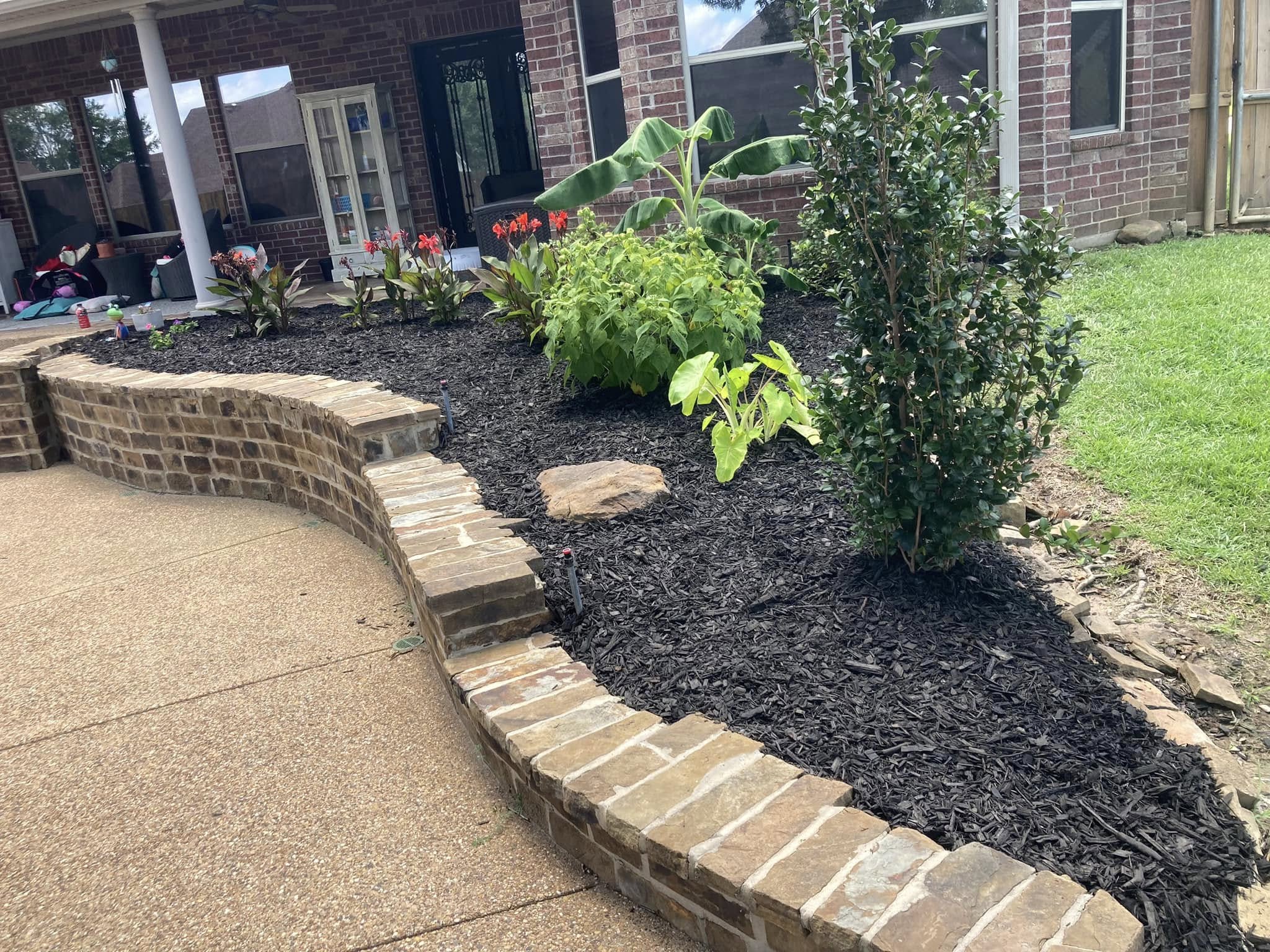 Curved natural-stone retaining wall with black mulch and shrubs along a brick home